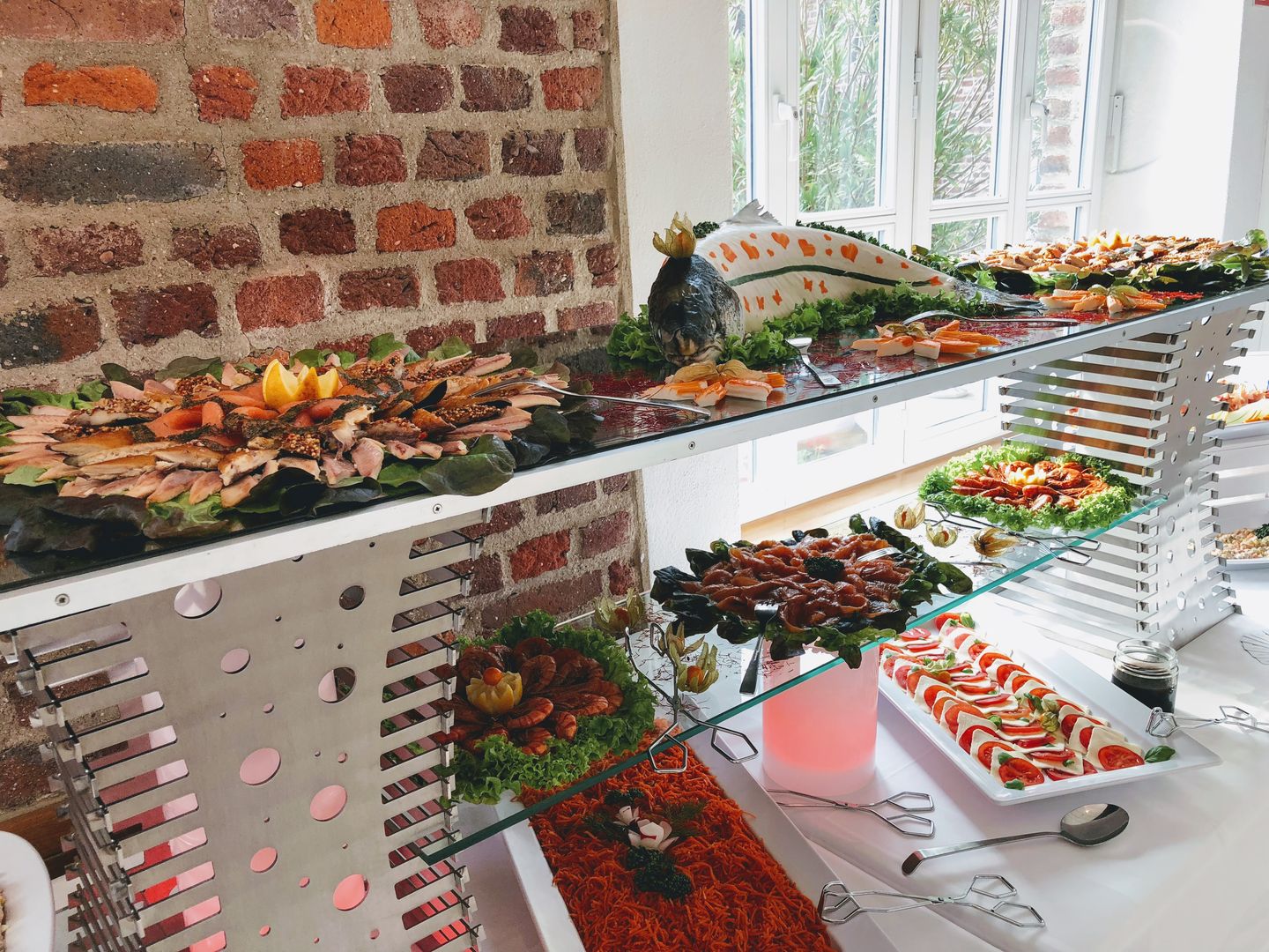 Fisch, kalte Platten und Salate auf Glasetageren vor einer Backsteinwand